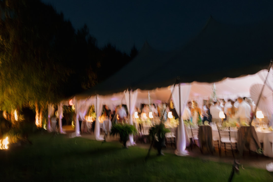 Blurry image of an outdoor evening event with a tent, warm lights, and guests mingling. A soft, festive atmosphere is suggested under a dark sky.