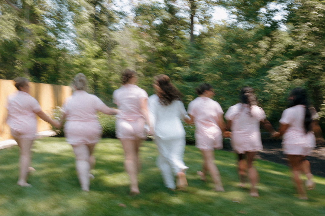 Bridesmaids running together across the lawn in matching outfits, captured candidly during getting ready.