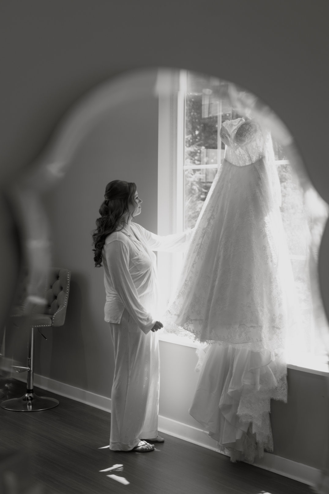 Black and white image of the bride standing near a window while her wedding dress hangs nearby, photographed by Chloe Elizabeth Photography.