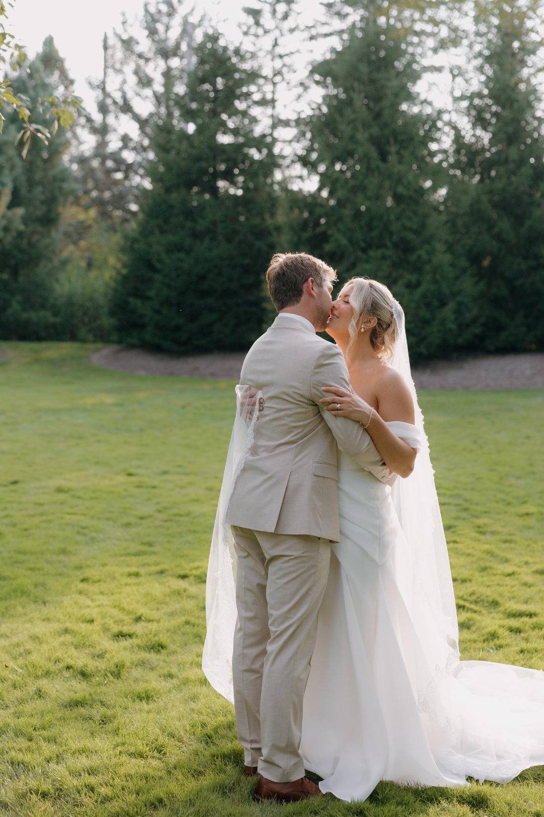 A couple embraces and kisses outdoors on a sunny day. The bride is in a white gown and veil, while the groom wears a light suit. Lush trees backdrop. Romantic atmosphere.