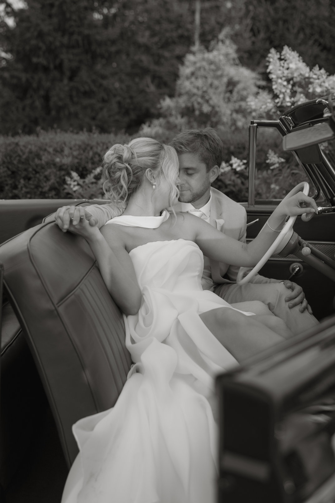 A black and white photo of a couple in wedding attire embracing in a classic convertible. The mood is romantic and serene, surrounded by lush greenery.