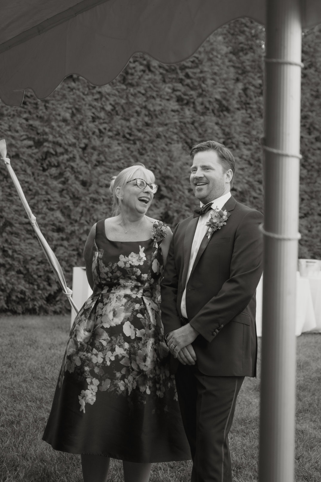 A woman in a floral dress and a man in a suit with a boutonniere joyfully laugh under a tent at an outdoor event, surrounded by lush greenery.