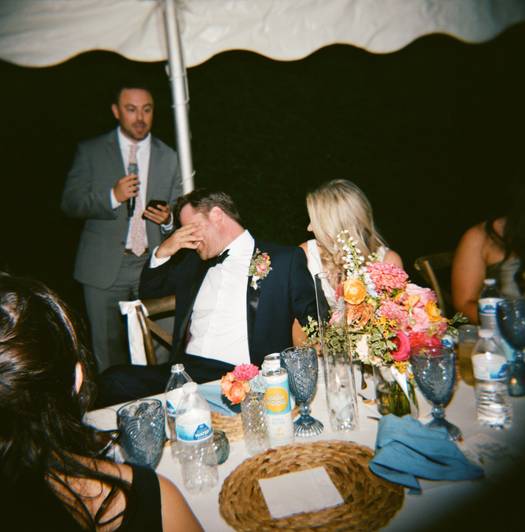 A man in a suit sits at a table, covering his face in laughter during a speech. A woman beside him smiles. Colorful floral arrangements and drinks adorn the table. The atmosphere is joyful and festive.