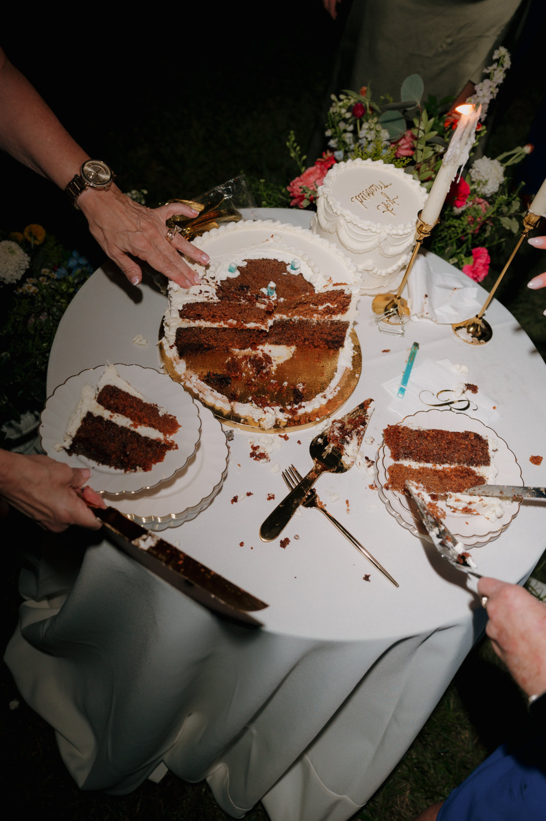 A table with a partially eaten cake, scattered slices, and crumbs. Hands with cutlery reach for more. Flowers and candles add a festive touch.