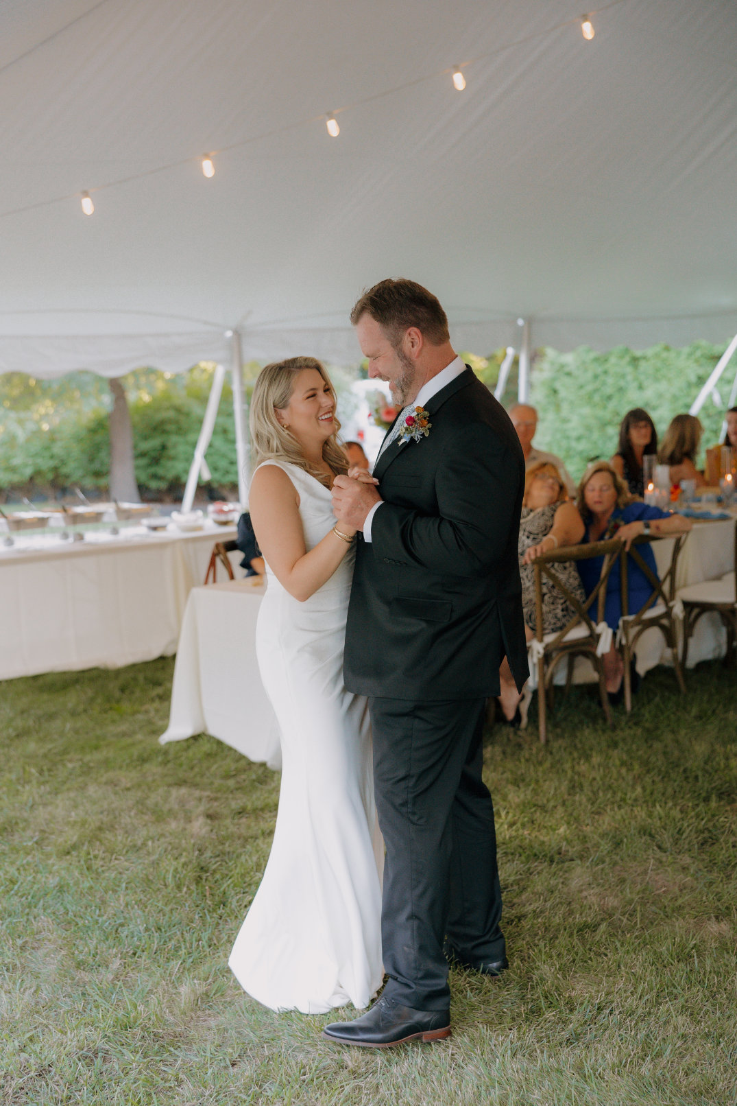 A couple shares a joyful dance at a wedding under a white tent adorned with string lights. Guests watch from rustic chairs, enhancing the festive ambiance.