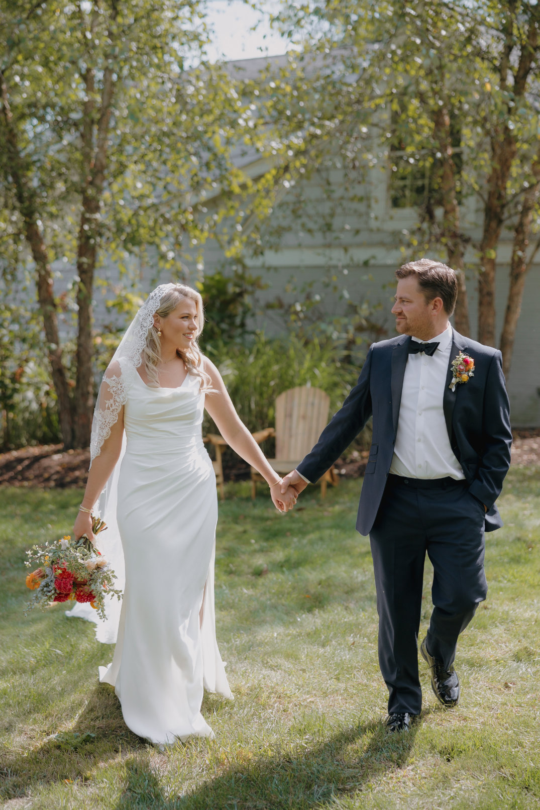 Bride in a white gown and lace veil holds a bouquet and groom's hand. Groom wears a navy suit. They smile while walking on grass by trees.