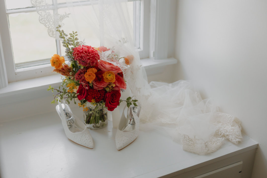 A vibrant bouquet of red, orange, and yellow flowers in a vase sits by white lace heels and a veil on a windowsill, conveying a bridal theme.