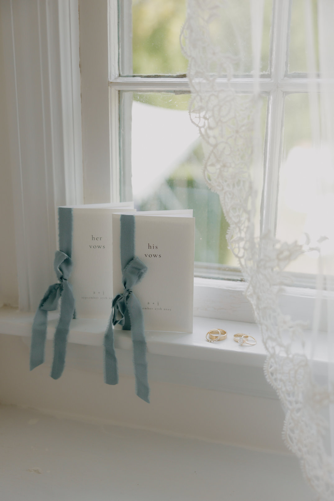 Two vow booklets with blue ribbons labeled "her vows" and "his vows" sit on a window sill beside two gold rings. Lace curtain partially visible.