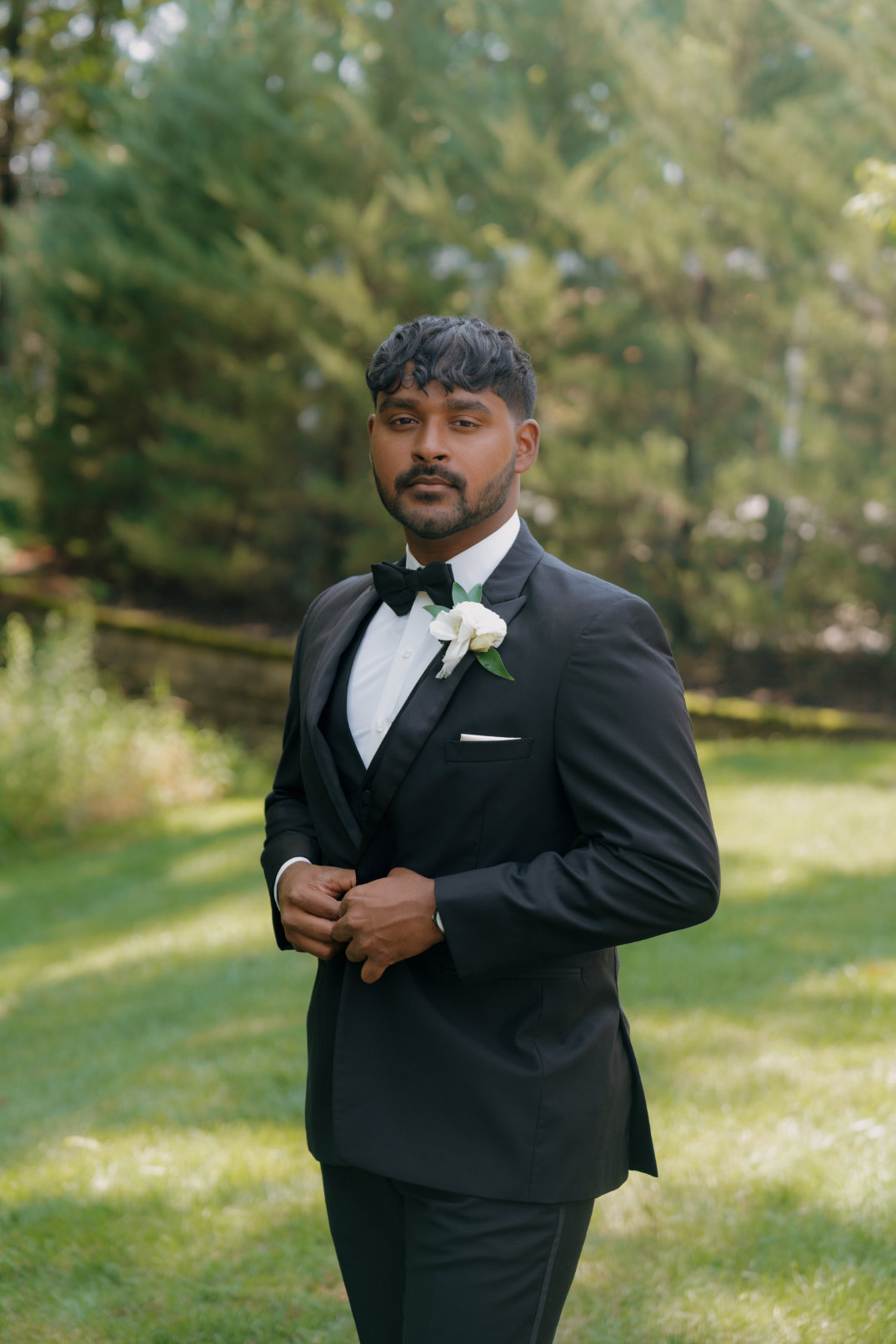 Groom standing outdoors in a tuxedo, adjusting his jacket and looking off-camera.