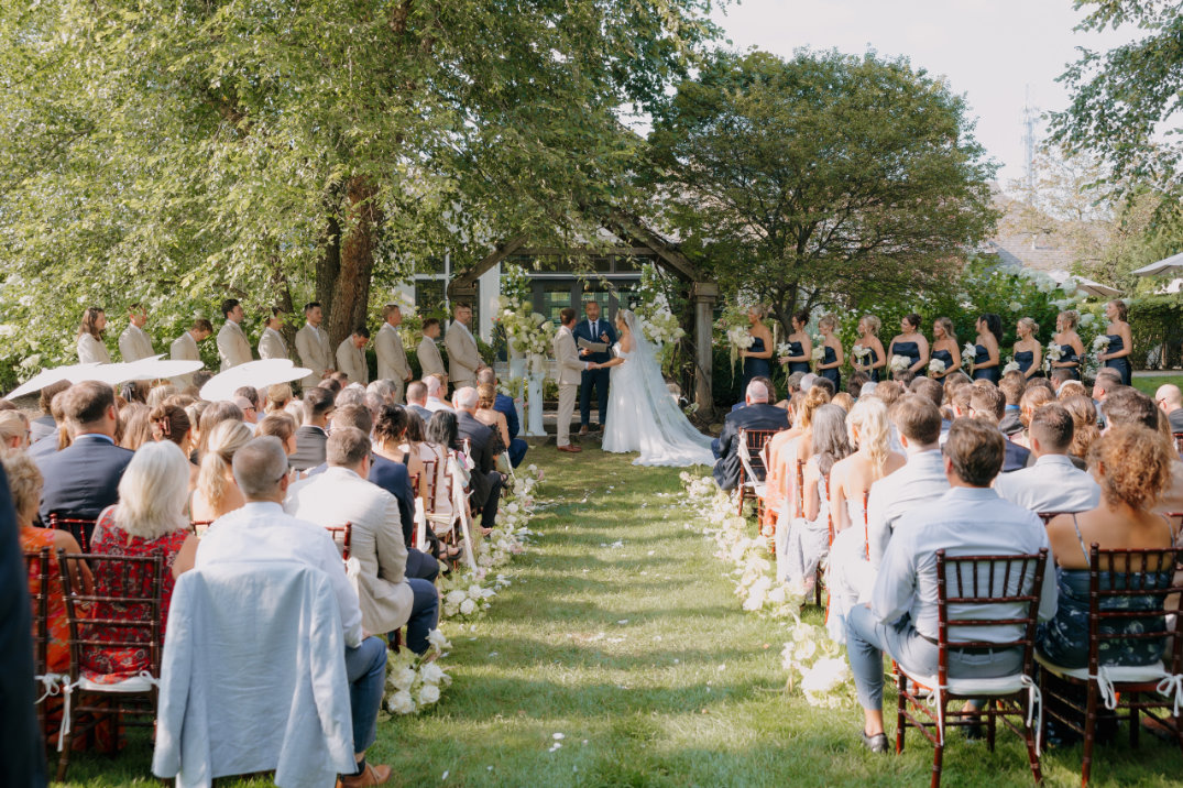 Outdoor wedding ceremony at The Orchid House Winery in Cleveland, Ohio, couple at altar, bridesmaids in dark dresses, groomsmen in beige suits, guests seated on wooden chairs.