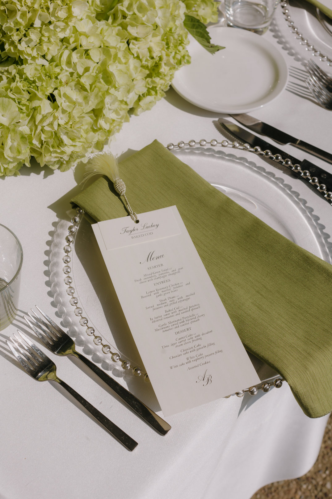 Wedding table setting with green hydrangeas, a beaded glass charger, a green napkin, and a menu card on white linens, conveying elegance and freshness.