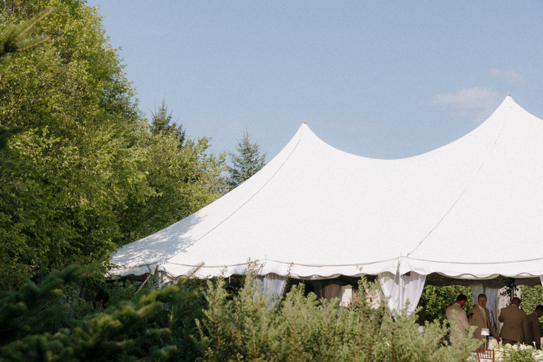 A large white tent set at Orchid House Wintery under a clear blue sky. People in formal attire gather inside, conveying a serene, celebratory atmosphere.