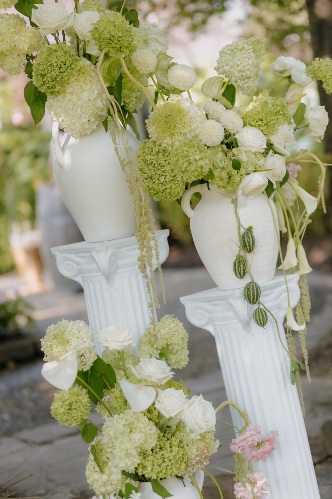 Elegant floral arrangement with white and green hydrangeas, roses, and greenery in white vases on classical columns. Serene outdoor setting.