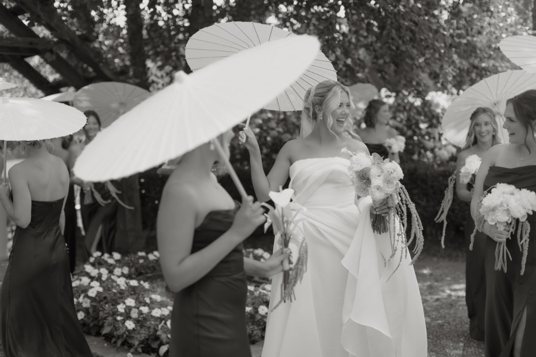 Black and white photo of a joyful bride and bridesmaids holding parasols and bouquets, standing in a garden. They wear elegant dresses and appear cheerful.