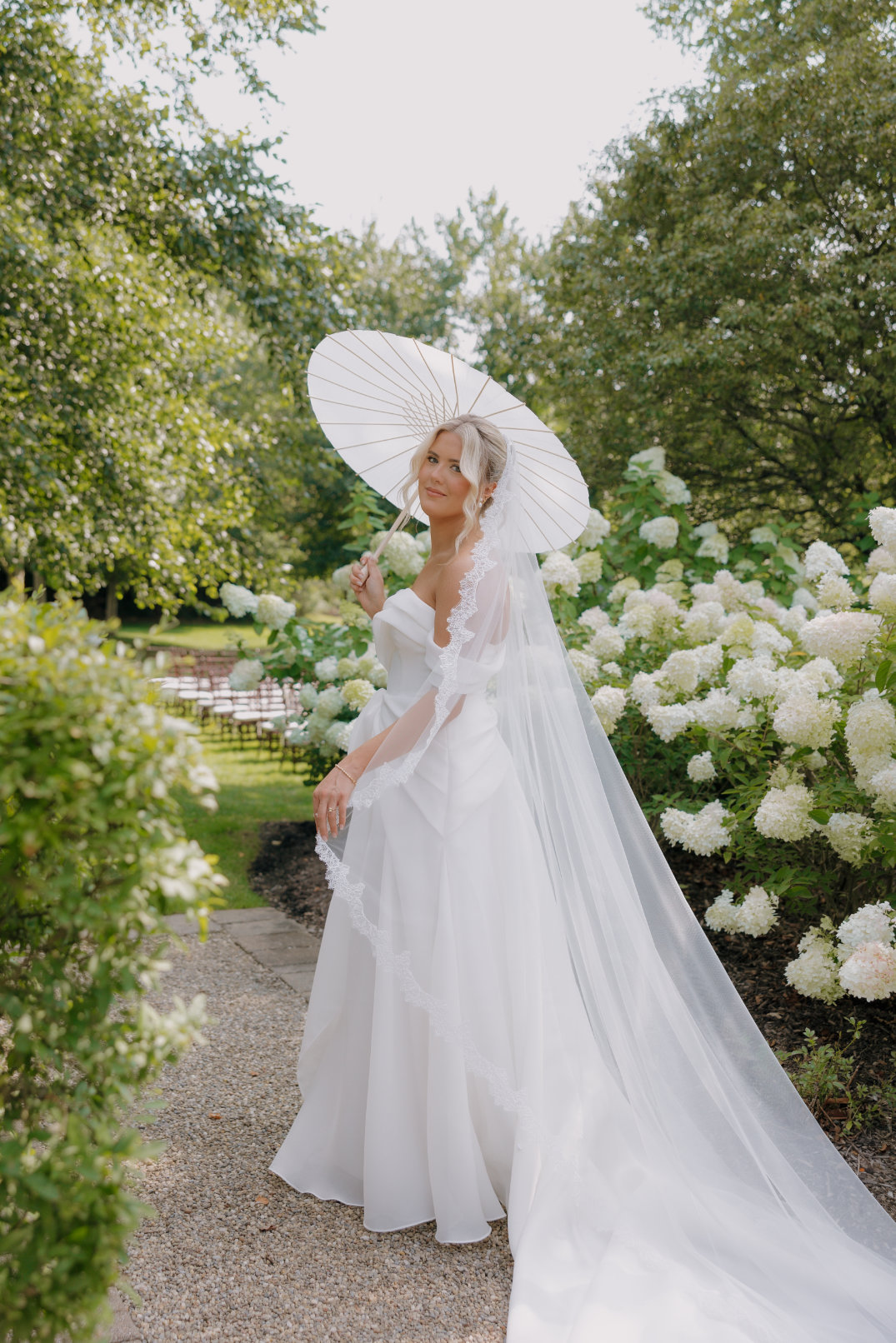 An Ohio bride in a flowing white gown holds a delicate parasol, standing on a garden path. She is surrounded by lush greenery and white hydrangeas, exuding a serene, elegant vibe.